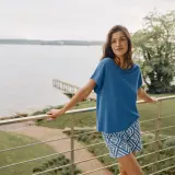 Frau auf Balkon mit Blick auf See, trägt blaues Oberteil aus modal stoff und gemusterte Shorts.