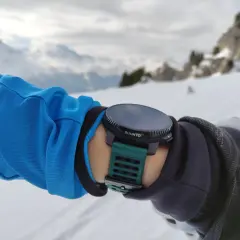 Suunto Vertical Test: Arm in blauer Jacke mit grüner Suunto-Uhr im Schnee, Bergkulisse.