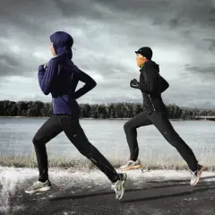 Zwei Läuferinnen in warmer Winterkleidung joggen am Fluss entlang. Die eine trägt einen blauen Hoodie und Hijab, die andere eine schwarze Jacke und orangefarbenen Schal.