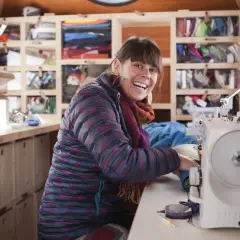 Frau lacht fröhlich an ihrer Nähmaschine, bereit für die Patagonia Reparatur. Regale voller bunter Outdoor-Kleidung im Hintergrund.