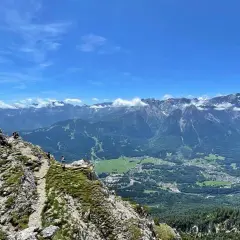 Gipfelkreuz auf dem Kramer­spitz, umgeben von Wanderern. Im Hintergrund das Alpenpanorama und ein Tal mit Häusern.