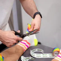 Hände schneiden buntes Kinesio-Tape zu. Die Frage "kinesio tape wann entfernen" wird hier visuell beantwortet: wenn es zugeschnitten wird, ist es bereit zur Anwendung.