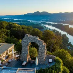 Der Rolandbogen thront über dem Rhein. Ein Restaurant mit Terrasse lädt zum Verweilen ein, während im Hintergrund Berge und ein Fluss im Abendlicht glänzen.