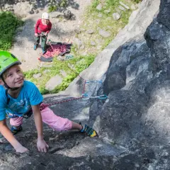 Ein Mädchen mit Helm klettert an einem Felsen, während eine andere Person sichert. Ein tolles Erlebnis beim Klettern mit Kindern.