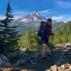 Frau mit Rucksack und Wanderstöcken auf einem steinigen Wanderweg in den USA, im Hintergrund ein schneebedeckter Berg und Nadelbäume.