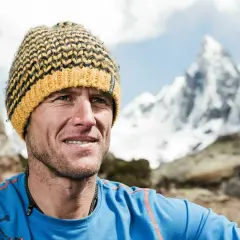 Stephan Siegrist, ein Bergsteiger, lächelt in die Kamera, eine gestreifte Mütze auf dem Kopf, vor einer schneebedeckten Bergkulisse.