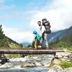 Vater und Sohn überqueren eine Brücke beim Wandern mit Kleinkind. Berge und ein Fluss im Hintergrund.