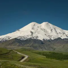 Der majestätische Elbrus Berg, bedeckt mit Schnee, ragt über grüne Hügel und eine kurvige Straße auf.
