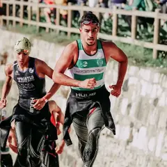 Zwei Athleten laufen bergauf, die Triathlon-Reihenfolge im Blick. Der vordere trägt ein grünes und weißes Shirt, der hintere ein schwarzes mit der Aufschrift "TRIATLO".