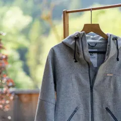 Graue Lodenjacke mit Kapuze, Reißverschluss und Taschen, aufgehängt an einem Holzständer vor blühendem Baum und grüner Natur.