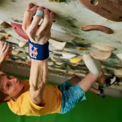 Alex Megos klettert konzentriert an einer Boulderwand. Seine Hand greift fest einen Griff, während sein Blick nach oben gerichtet ist.