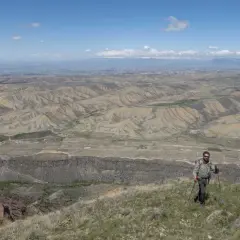 Ein Mann mit Rucksack und Wanderstöcken genießt die weite Aussicht bei seinem Armenien Wandern. Die Landschaft ist geprägt von trockenen Hügeln und einer fernen Stadt.