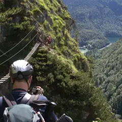 Ein Mann mit Helm und Rucksack fotografiert einen Klettersteig in Berchtesgaden. Eine Hängebrücke führt über eine Schlucht mit Blick auf einen See.