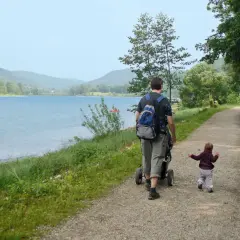 Vater schiebt Kinderwagen am Happurger Stausee entlang, während das Kind vorausläuft. Herrliche Kulisse für eine Wanderung.