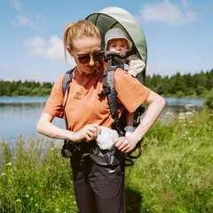Frau mit kraxe baby am Seeufer, umgeben von grüner Natur und Bäumen.