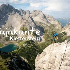 Ein Kletterer sichert sich am Tajakante Klettersteig mit Blick auf einen Bergsee.