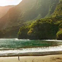 Sonne beleuchtet den malerischen **Madeira schwarzer Strand** mit üppig grünen Bergen im Hintergrund. Wellen brechen sanft am Ufer.