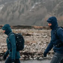 Zwei Wanderer mit Rucksäcken gehen durch eine steinige Landschaft in Schottland. Das Wetter ist regnerisch, die Kapuzen sind hochgezogen.