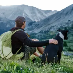 Mann mit deuter Rucksack und Hund sitzt auf Bergwiese.
