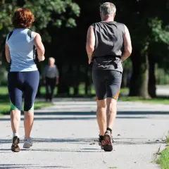 Ein Paar genießt **slow jogging** auf einem Weg im Park. Im Hintergrund eine weitere Person.