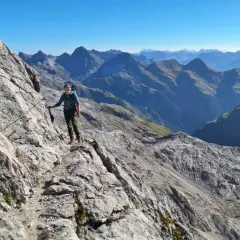 Frau auf dem **Heilbronner Weg wandern**, gesichert an einem Stahlseil, mit atemberaubendem Bergpanorama im Hintergrund.