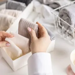 Hände sortieren gefaltete Kleidung, darunter auch warme **Wandersocken**, in einem Behälter. Daneben ein Korb mit weiteren Textilien und eine Kaffeetasse.