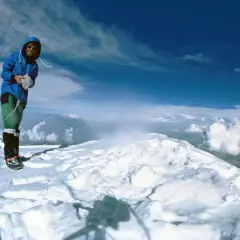Reinhard Messner steht auf einem schneebedeckten Gipfel, umgeben von Wolken und blauem Himmel.