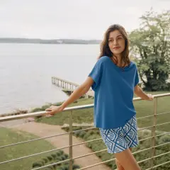 Frau auf Balkon mit Blick auf See, trägt blaues Oberteil aus modal stoff und gemusterte Shorts.