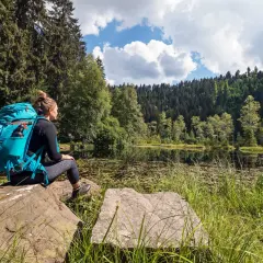 Frau mit Rucksack sitzt an einem See, umgeben von Wald. Ein perfekter Moment auf einer der vielen **weitwanderwege europa**.