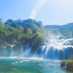 Ein majestätischer Krk Wasserfall stürzt über mehrere Stufen in ein türkisfarbenes Becken. Üppige grüne Bäume säumen die Klippen und der Himmel ist strahlend blau.