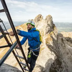 Mann klettert auf einem Leiternabschnitt des Oberstdorf Klettersteigs. Die Aussicht auf die Alpen ist atemberaubend.