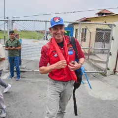Kenton Cool, lächelnd, trägt eine rote Jacke und einen blauen Hut. Er steht vor einem Tor, umgeben von Kameraleuten.