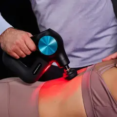 Eine Hand führt eine Massagepistole zur Anwendung am unteren Rücken einer Person. Rotes Licht leuchtet auf.