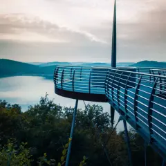 Moderne Aussichtsplattform über einem See, ideal für das Wandern in NRW. Sanfte Hügel und ein dramatischer Himmel im Hintergrund.