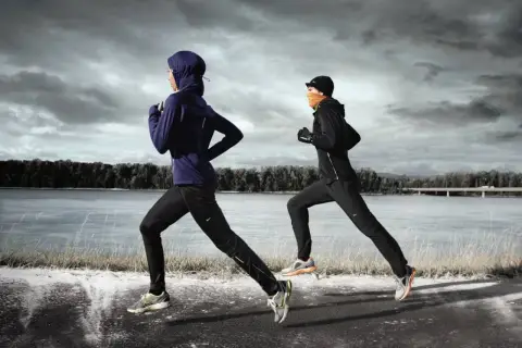 Zwei Läuferinnen in warmer Winterkleidung joggen am Fluss entlang. Die eine trägt einen blauen Hoodie und Hijab, die andere eine schwarze Jacke und orangefarbenen Schal.