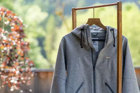 Graue Lodenjacke mit Kapuze, Reißverschluss und Taschen, aufgehängt an einem Holzständer vor blühendem Baum und grüner Natur.