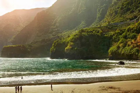 Sonne beleuchtet den malerischen **Madeira schwarzer Strand** mit üppig grünen Bergen im Hintergrund. Wellen brechen sanft am Ufer.