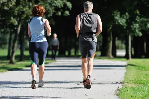 Ein Paar genießt **slow jogging** auf einem Weg im Park. Im Hintergrund eine weitere Person.