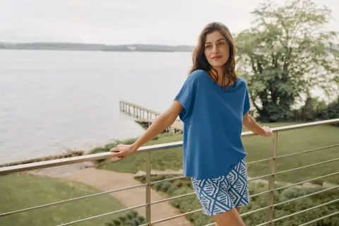 Frau auf Balkon mit Blick auf See, trägt blaues Oberteil aus modal stoff und gemusterte Shorts.