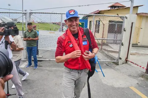 Kenton Cool, lächelnd, trägt eine rote Jacke und einen blauen Hut. Er steht vor einem Tor, umgeben von Kameraleuten.