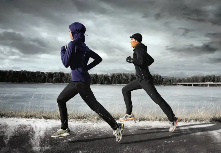 Zwei Läuferinnen in warmer Winterkleidung joggen am Fluss entlang. Die eine trägt einen blauen Hoodie und Hijab, die andere eine schwarze Jacke und orangefarbenen Schal.
