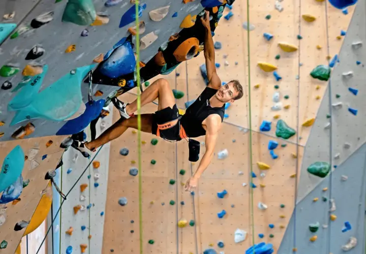 Yannick Flohé klettert an einer Boulderwand, hält sich an einem bunten Griff fest und blickt lächelnd in die Kamera.