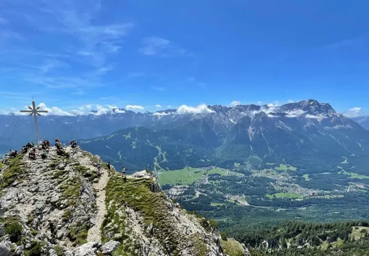 Gipfelkreuz auf dem Kramer­spitz, umgeben von Wanderern. Im Hintergrund das Alpenpanorama und ein Tal mit Häusern.