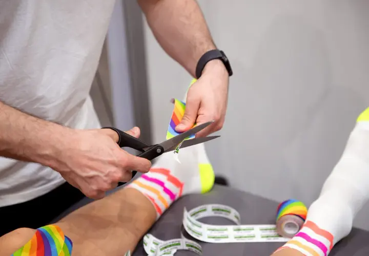 Hände schneiden buntes Kinesio-Tape zu. Die Frage "kinesio tape wann entfernen" wird hier visuell beantwortet: wenn es zugeschnitten wird, ist es bereit zur Anwendung.