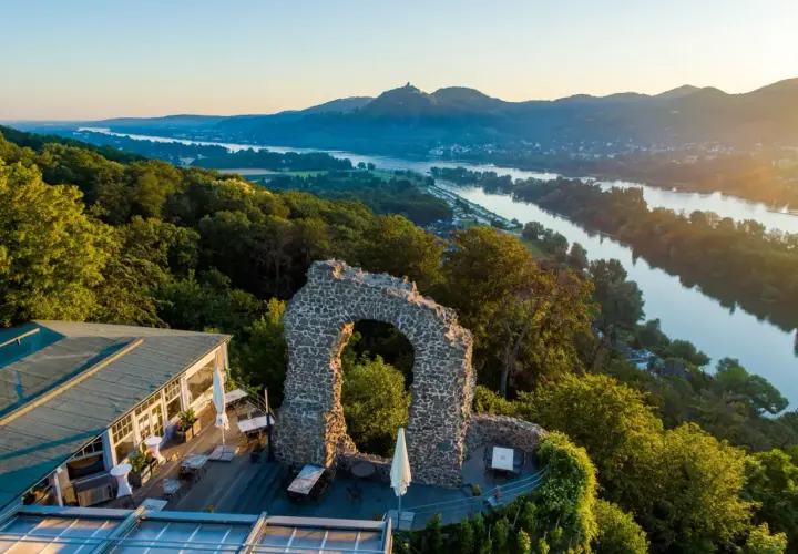 Der Rolandbogen thront über dem Rhein. Ein Restaurant mit Terrasse lädt zum Verweilen ein, während im Hintergrund Berge und ein Fluss im Abendlicht glänzen.