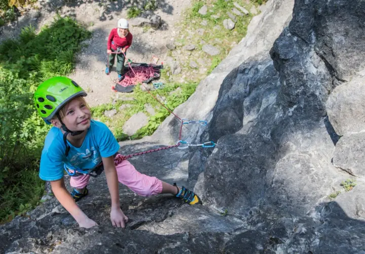 Ein Mädchen mit Helm klettert an einem Felsen, während eine andere Person sichert. Ein tolles Erlebnis beim Klettern mit Kindern.