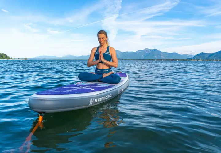 Frau praktiziert SUP Yoga auf einem Brett im See, mit Bergen im Hintergrund.