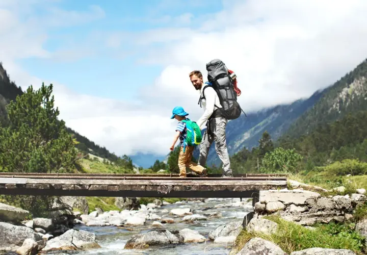Vater und Sohn überqueren eine Brücke beim Wandern mit Kleinkind. Berge und ein Fluss im Hintergrund.