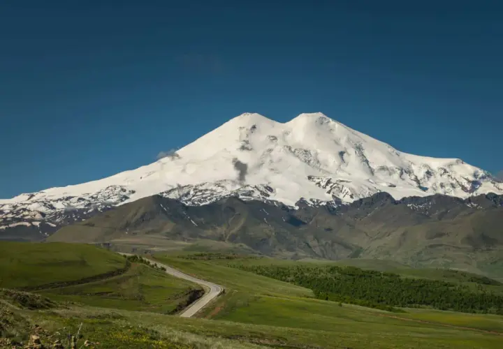Der majestätische Elbrus Berg, bedeckt mit Schnee, ragt über grüne Hügel und eine kurvige Straße auf.