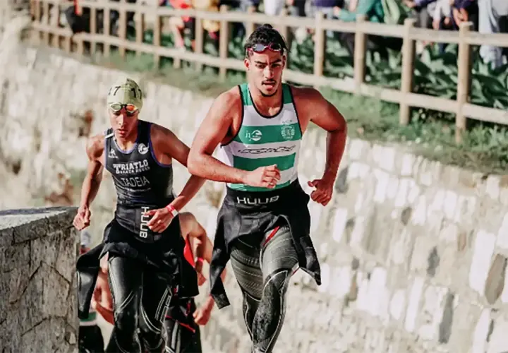 Zwei Athleten laufen bergauf, die Triathlon-Reihenfolge im Blick. Der vordere trägt ein grünes und weißes Shirt, der hintere ein schwarzes mit der Aufschrift "TRIATLO".